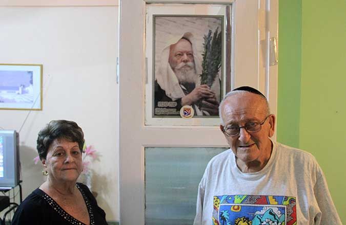 Rebeca and Alberto Meshulam, 70 and 78, respectively, in front of a portrait of the late Lubavitcher rebbe, Menachem Mendel Schneerson. The Meshulams host Lubavitch emissaries at their Havana apartment throughout the year. (Josh Tapper)